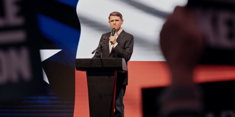 A man in a suit speaks at a podium on stage, with a large Texas flag projected behind him and blurred audience signs in the foreground.