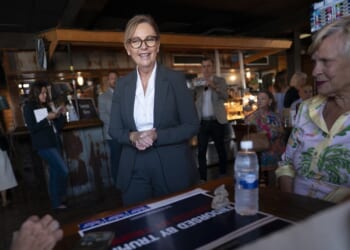 A woman in a suit smiles while standing near a table with a campaign sign and water bottle. People sit and mingle in a casual indoor setting with warm lighting.