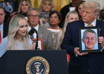 A woman Anne Fundner speaks at a podium while President Trump stands next to her and a framed photo of a young person, her son; people in the background also hold photos.