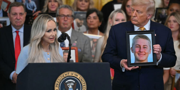 A woman Anne Fundner speaks at a podium while President Trump stands next to her and a framed photo of a young person, her son; people in the background also hold photos.