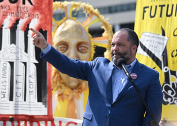 A new report highlights foreign funding for climate activist groups. Pictured: Sierra Club Executive Director Ben Jealous, a man in a blue suit speaks into a microphone and points, with climate protest signs and a sun mask in the background. Signs criticize banks and call for action against climate change.