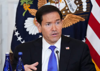 Marco Rubio in a suit speaks at a table with two blue bottles in front of him. Behind him are the U.S. flag and the presidential seal.