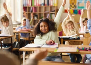 Children in a classroom sit with their hands raised toward the teacher.