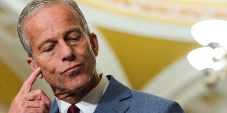 A man in a blue suit touches his ear and looks down pensively, standing indoors with yellow and white decor in the background.