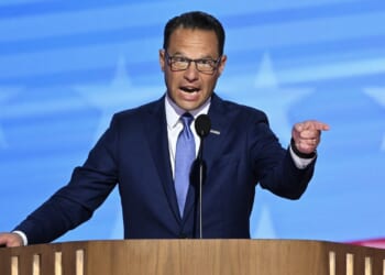 A man in a blue suit and tie speaks passionately at a wooden podium, pointing with his right hand. The background features a blue pattern with white stars, resembling the American flag.