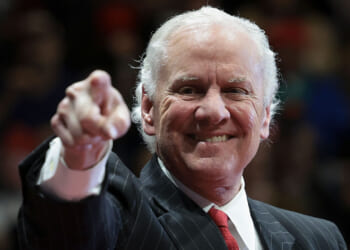 An older man in a suit and red tie smiles while pointing toward the camera. The background is blurred, focusing attention on his expression and gesture.