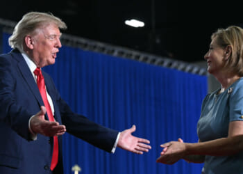 Donald Trump, in a suit with a red tie, extends his arms toward Darline Graham in a blue dress, both smiling and standing in front of blue curtains and an American flag.
