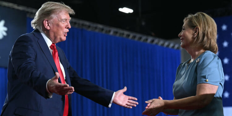 Donald Trump, in a suit with a red tie, extends his arms toward Darline Graham in a blue dress, both smiling and standing in front of blue curtains and an American flag.