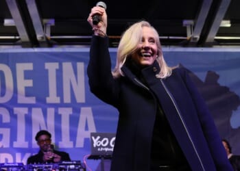 Abigail Spanberger with blonde hair smiles and raises a microphone on stage. A musician performs on equipment behind her. The background features a blue banner with partially visible text.
