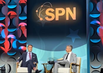 Former Virginia Attorney General Jason Miyares sits on stage alongside State Policy Network's Tony Woodlief under a large SPN sign, engaged in conversation. The backdrop features a blue and red geometric pattern and spotlights. A small table with water glasses is between them.