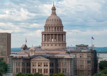 The Texas State Capitol building in Austin, with scaffolding on parts of its exterior, stands under a cloudy sky. The Texas flag is visible on the right side.