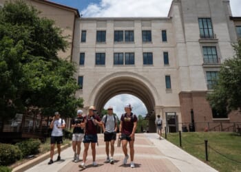 Six people walk on a pathway outside a large, beige university building with an archway, surrounded by trees under a partly cloudy sky.