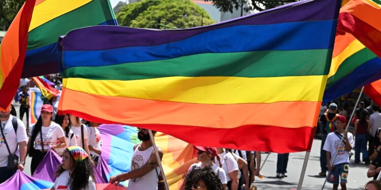 People march in a parade holding rainbow pride flags. Many wear white shirts and colorful accessories, celebrating LGBTQ+ pride outdoors under trees and sunlight.