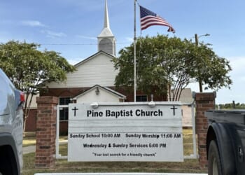 White church with a steeple and American flag, seen behind a sign reading Pine Baptist Church with service times, flanked by parked vehicles on grass. Trees frame the church against a blue sky.