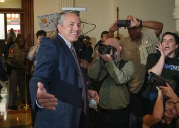 A man in a suit smiles and gestures while surrounded by photographers and people, some with cameras and others reacting energetically, at an indoor event.