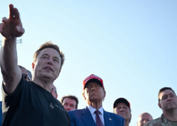 Elon Musk in a black shirt gestures while speaking to Donald Trump and a group of people outdoors.