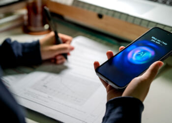 A person writes on paper while holding a smartphone displaying a digital assistant with What can I help you with? and AI on the screen, illustrating how technology reduces friction in daily tasks. A laptop and office supplies are on the desk in the background.