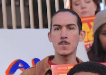 A person with a mustache stands in front of a white protest sign, holding a red pamphlet. Others are visible in the background, including a woman in a beanie. The mood appears serious and focused.