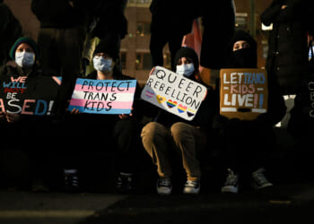 The transgender movement's arguments in court have changed from aggressive promotion to defense. Pictured: Five people sitting, holding protest signs with messages supporting trans and queer rights, such as Protect Trans Kids and Let Trans Kids Live. All are wearing masks, and their faces are partially obscured by shadows.