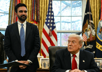 A man in a suit stands next to another man sitting at a desk in an office with U.S. flags, curtains, and framed photos in the background.
