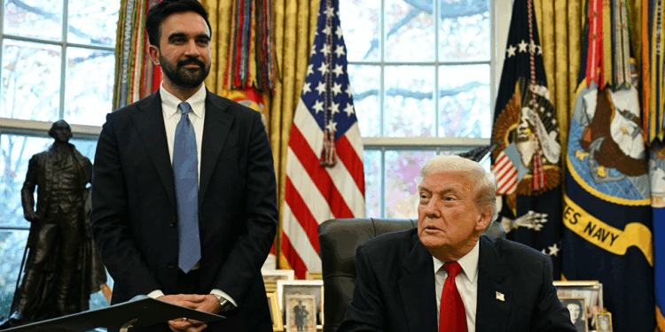 A man in a suit stands next to another man sitting at a desk in an office with U.S. flags, curtains, and framed photos in the background.