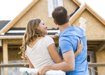Homeownership concept. Pictured: a couple stands in front of a partially built house, embracing. The man points toward the home while the woman holds rolled blueprints, both looking at the construction with anticipation.