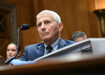 Dr. Anthony Fauci in a suit sits at a desk with a microphone, appearing serious, in a formal wooden room. Other people are seated in the background.