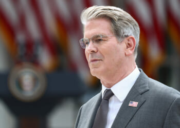 Scott Bessent in a suit with an American flag pin stands outdoors, with blurred U.S. flags and a presidential podium visible in the background.