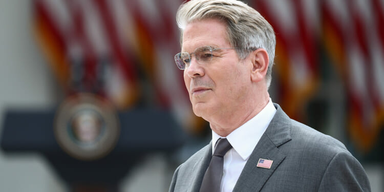 Scott Bessent in a suit with an American flag pin stands outdoors, with blurred U.S. flags and a presidential podium visible in the background.