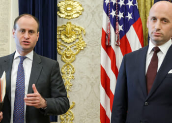 Two men in suits stand in a formal room with US flags and gold decorations behind them. One man holds papers and gestures while the other stands with a neutral expression.