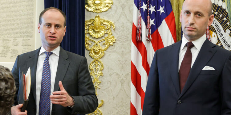 Two men in suits stand in a formal room with US flags and gold decorations behind them. One man holds papers and gestures while the other stands with a neutral expression.