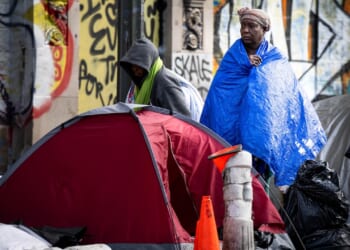 Two people stand near a red tent among belongings on a city street, wrapped in blankets, with graffiti on the wall behind them and bags and a traffic cone nearby.