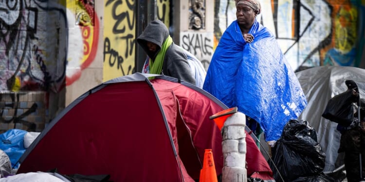 Two people stand near a red tent among belongings on a city street, wrapped in blankets, with graffiti on the wall behind them and bags and a traffic cone nearby.