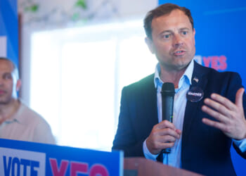 Tom Perriello speaks into a microphone at a podium with VOTE YES signs, while Hakeem Jeffries stands in the background.
