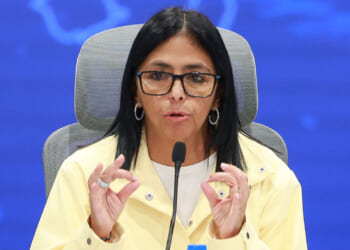 Delcy Rodriguez with long dark hair and glasses speaks into a microphone at a conference. She wears a yellow jacket and white shirt, gesturing with both hands. The background is blue.