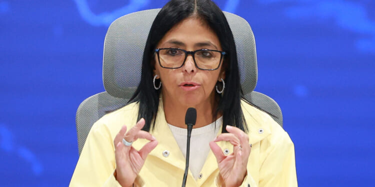 Delcy Rodriguez with long dark hair and glasses speaks into a microphone at a conference. She wears a yellow jacket and white shirt, gesturing with both hands. The background is blue.
