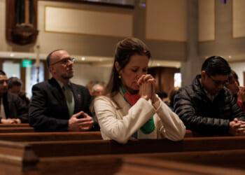 A group of people sits in church pews, heads bowed and hands clasped in prayer, in a softly lit sanctuary.