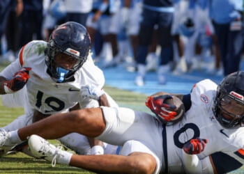 Two football players in white Virginia uniforms are on the field; one is holding the ball while laying down, and the other is behind him, with a blurred crowd and players in blue in the background.