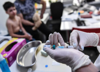 A healthcare worker prepares a measles vaccine injection in a clinic, while a shirtless child holding a teddy bear sits nearby with an adult. Medical supplies are on the table.