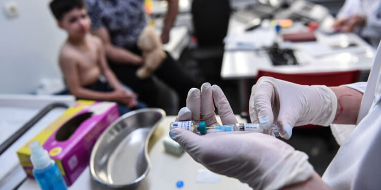 A healthcare worker prepares a measles vaccine injection in a clinic, while a shirtless child holding a teddy bear sits nearby with an adult. Medical supplies are on the table.