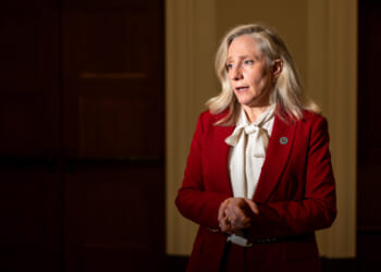 Virginia Gov. Abigail Spanberger, who backed the redistricting effort. Pictured: a woman with long blonde hair, wearing a red blazer and white blouse, stands indoors with hands clasped, looking concerned. The background is dimly lit with wooden doors.