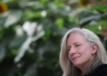 Abigail Spanberger with long gray hair smiles softly, sitting outdoors in Virginia with blurred green foliage and sunflowers in the background.