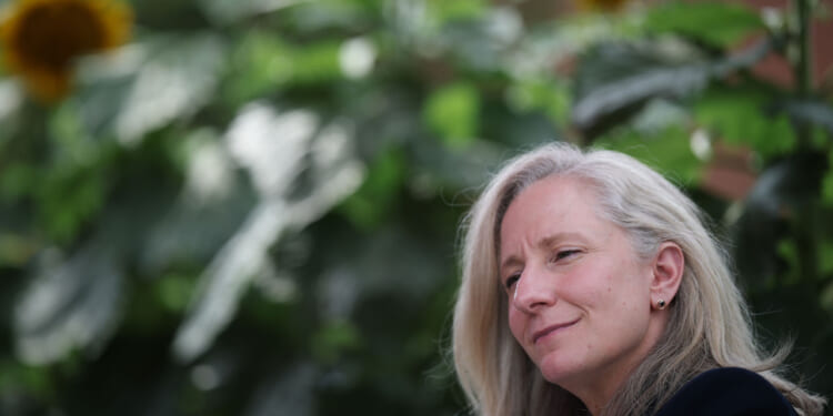 Abigail Spanberger with long gray hair smiles softly, sitting outdoors in Virginia with blurred green foliage and sunflowers in the background.
