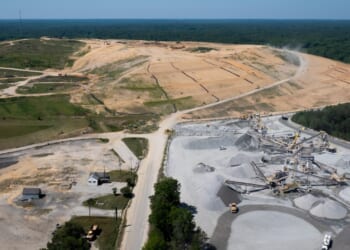 Aerial view of a landfill construction site with cleared land, earth-moving machinery, gravel piles, and a few small buildings, surrounded by trees and forested areas.