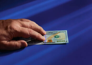 A vigilant hand holding a $100 US dollar bill on a smooth blue surface with light streaks casting shadows.