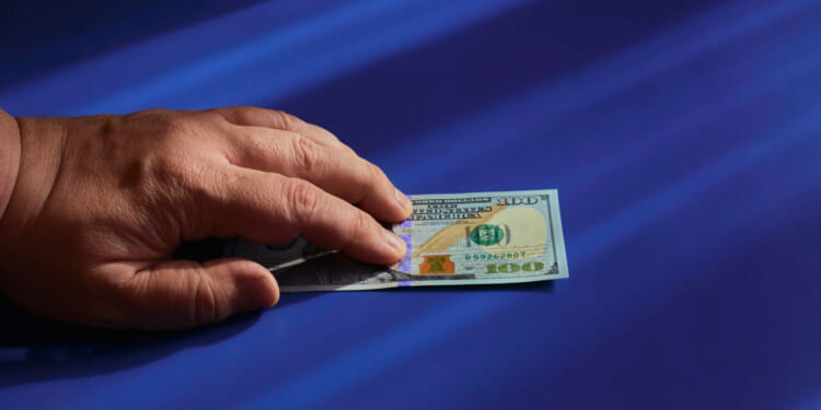 A vigilant hand holding a $100 US dollar bill on a smooth blue surface with light streaks casting shadows.