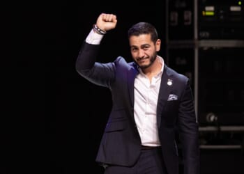 A man in a dark suit stands on stage with his right fist raised and a slight smile, in front of a dark background with electronic equipment visible.
