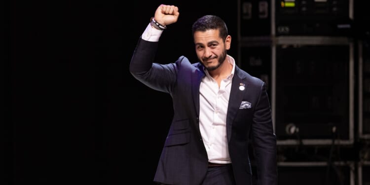 A man in a dark suit stands on stage with his right fist raised and a slight smile, in front of a dark background with electronic equipment visible.