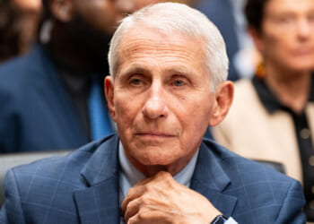 An older man with short white hair in a blue suit sits thoughtfully with his hand on his chin at what appears to be a formal hearing, with blurred people in the background.
