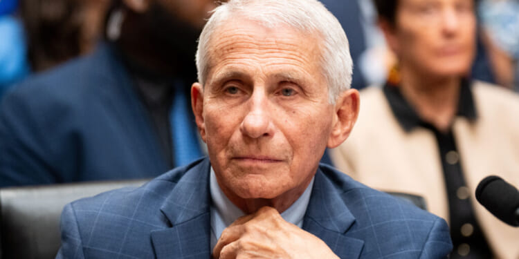 An older man with short white hair in a blue suit sits thoughtfully with his hand on his chin at what appears to be a formal hearing, with blurred people in the background.
